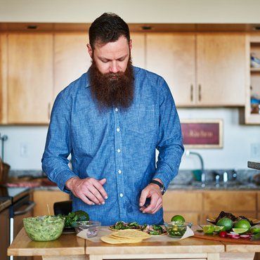 Bearded man in a blue shirt plating tacos in a kitchen