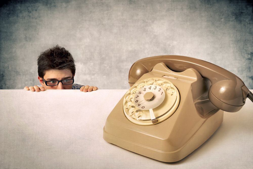 Man in glasses peeking nervously over a wall at a vintage telephone